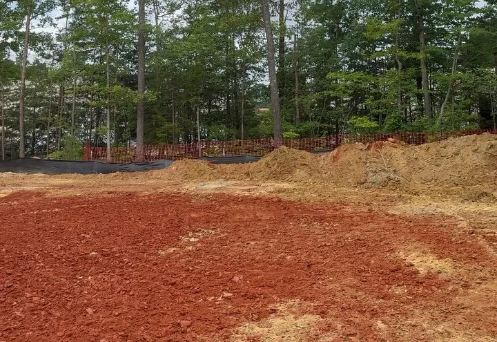 Cut red clay lot with silt fence and orange safety fence along the tree line and a spoil pile behind it