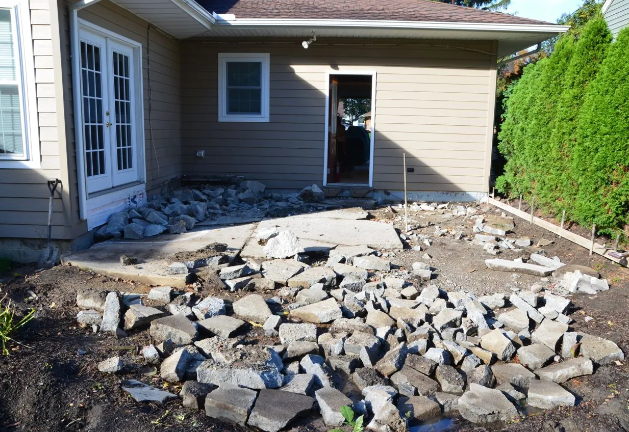Broken pieces of an old concrete patio spread across the ground behind a house