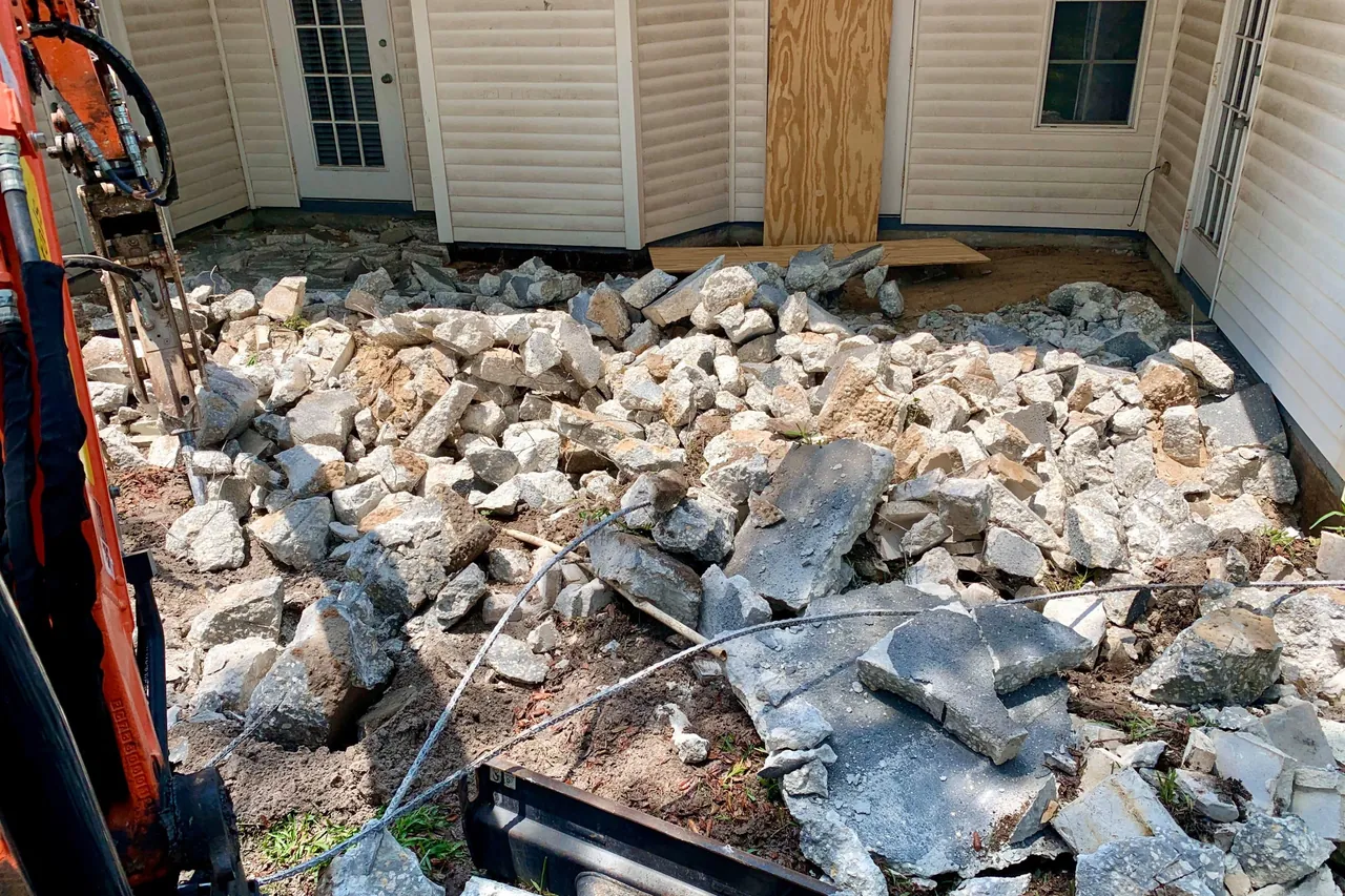 Broken concrete from a removed slab piled against the back of a house, with an excavator arm at the edge of the frame