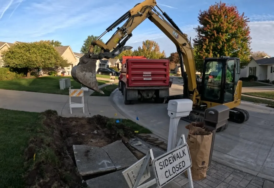 Mini excavator lifting out a broken concrete sidewalk beside a residential street, with a dump truck and a sidewalk closed sign
