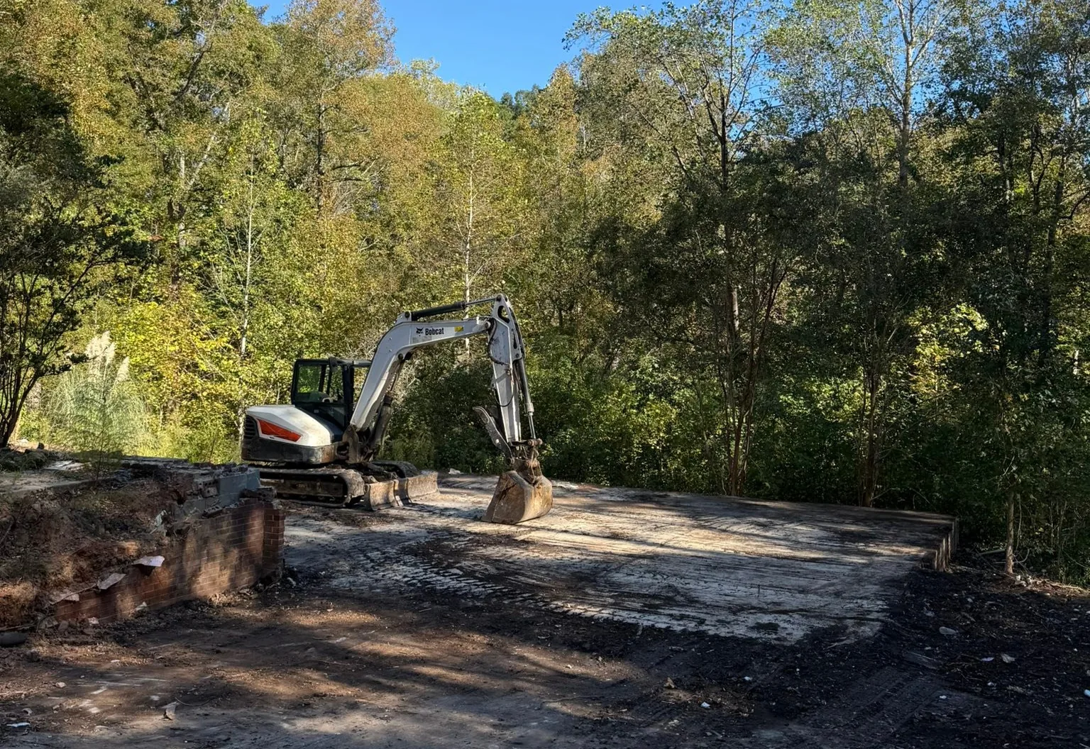 Excavator parked on a stripped pad where a structure stood, old brick foundation remnants at the edge