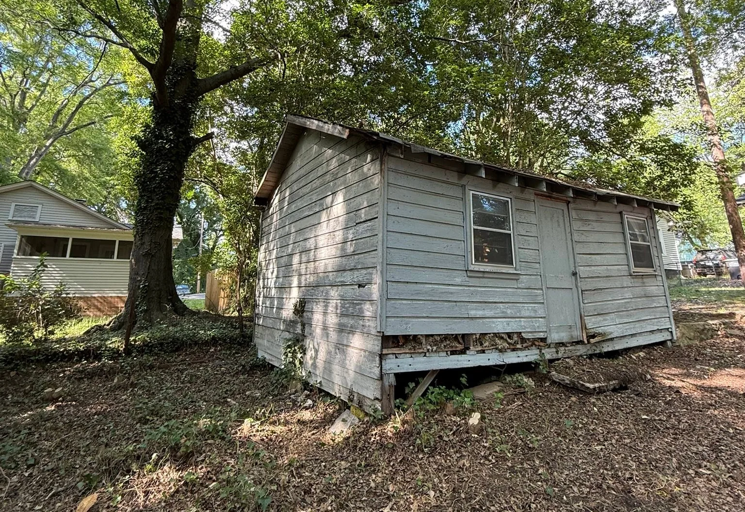 Old wood-sided outbuilding with a sagging floor under mature trees, before a teardown