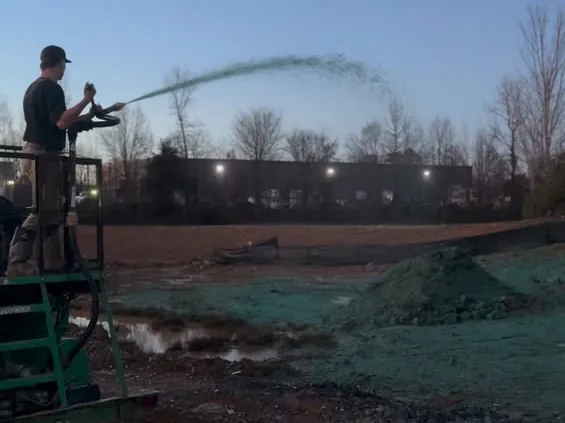 Operator spraying green seed and mulch over a graded pad at dusk with a silt fence line behind it