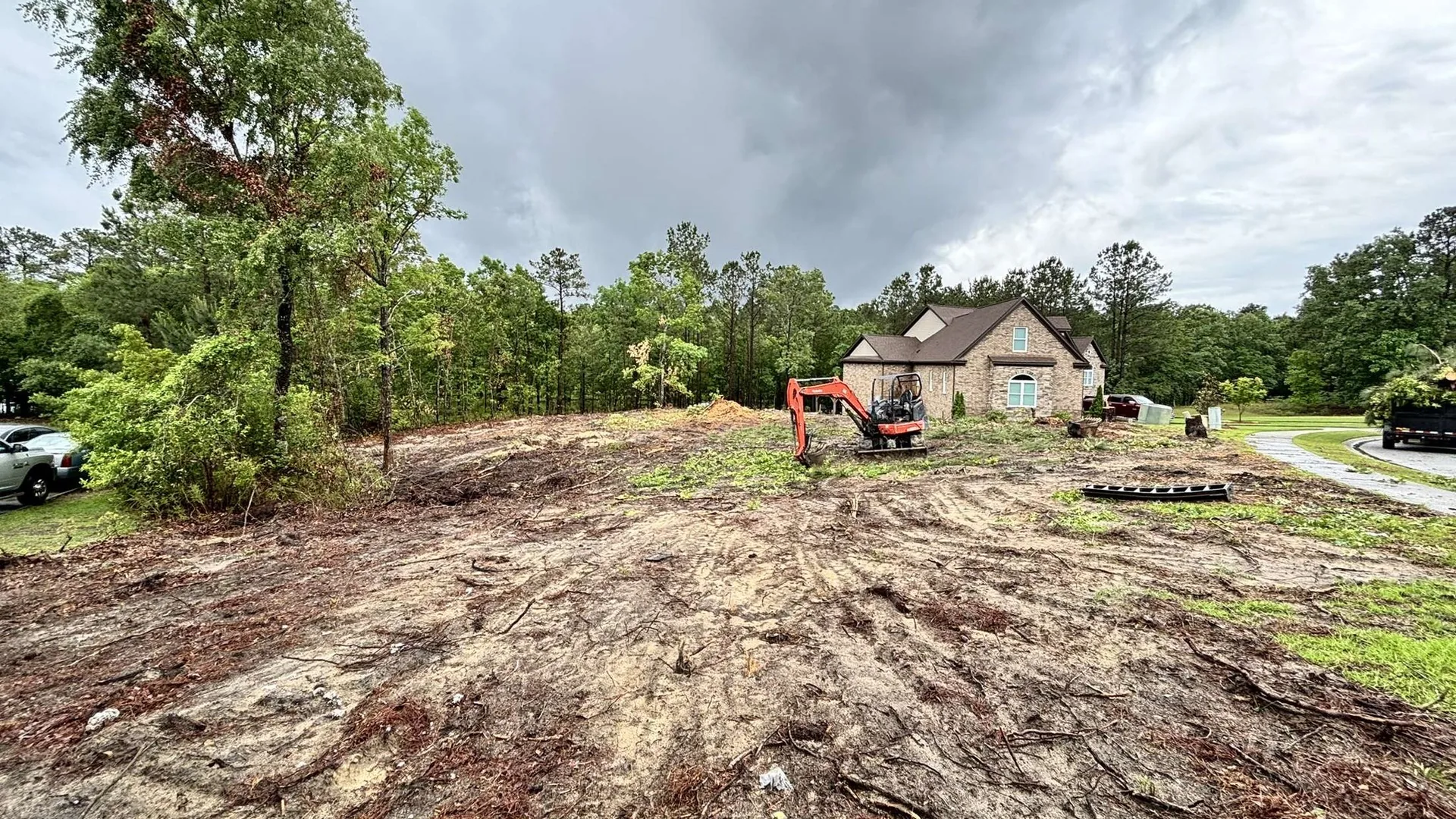 Mini excavator on a cleared lot beside a large brick house under a storm sky