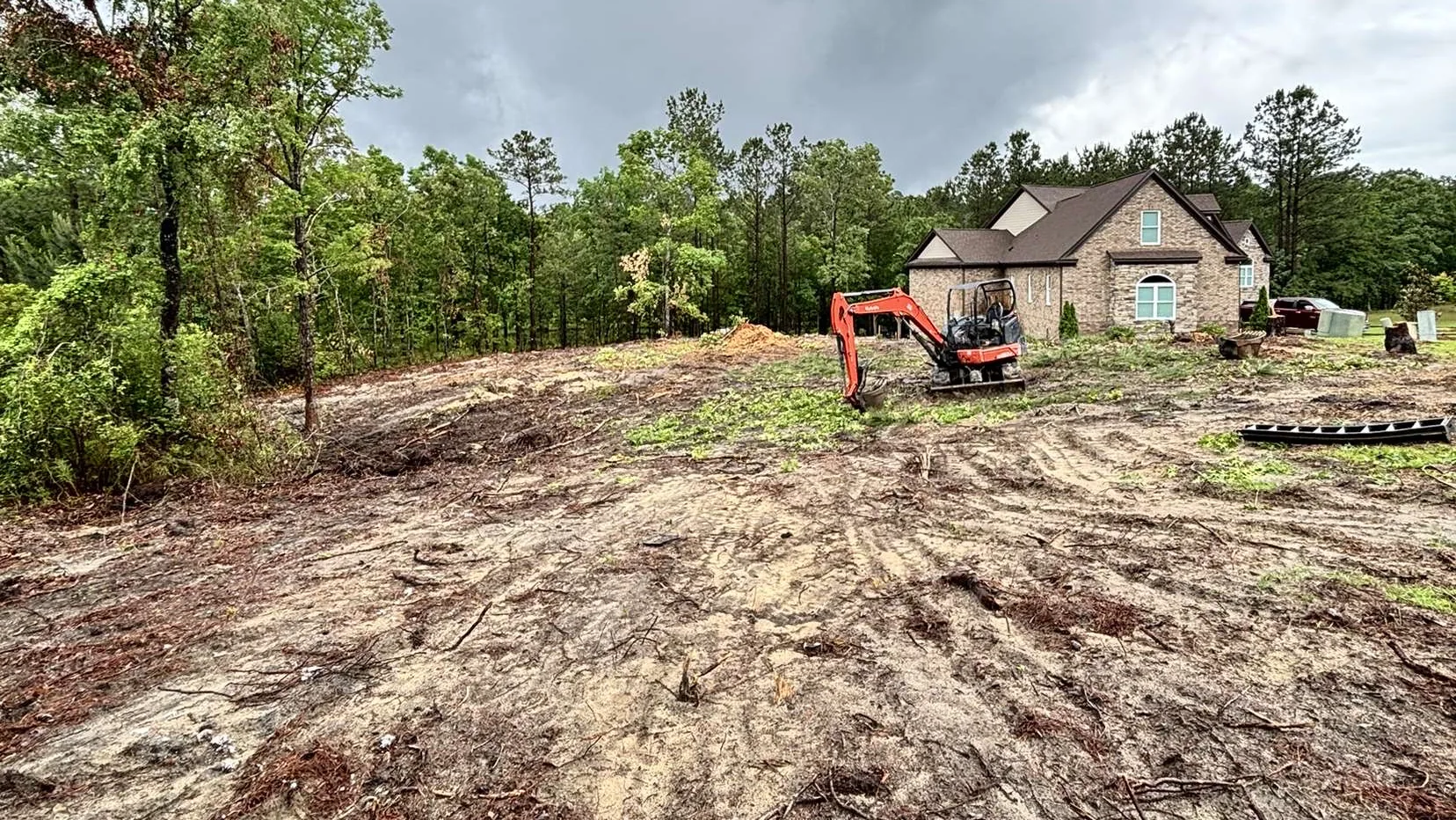 Mini excavator working a cleared lot of red clay and wood chips beside a new brick home