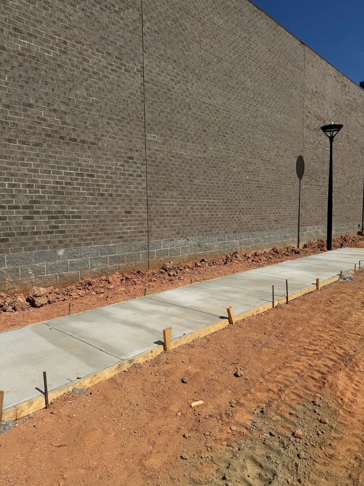 Red clay backfilled along a brick building's block foundation course beside a new concrete sidewalk