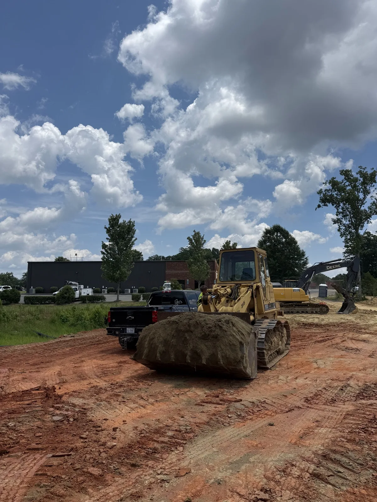 Track loader carrying a full bucket of red clay across a site with an excavator working behind it