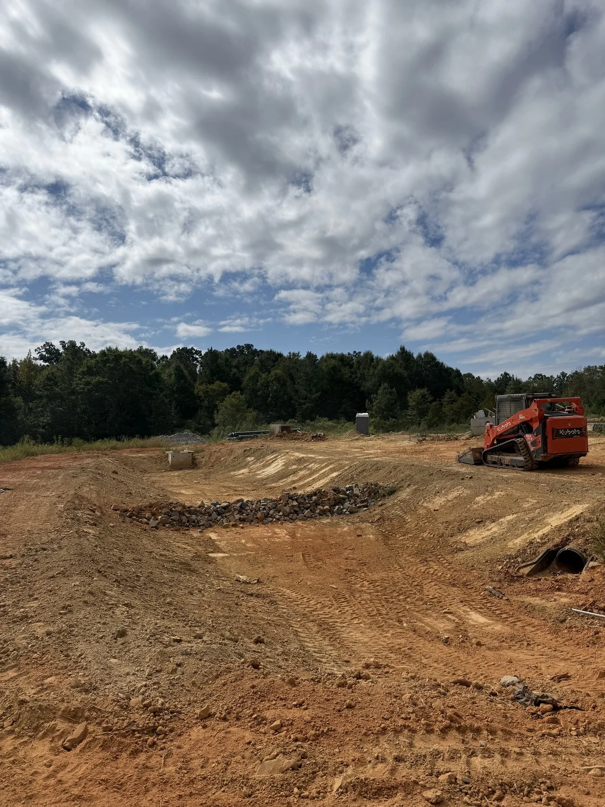 Compact track loader on a graded red clay site beside a pile of excavated rock, with a section of culvert pipe in the foreground