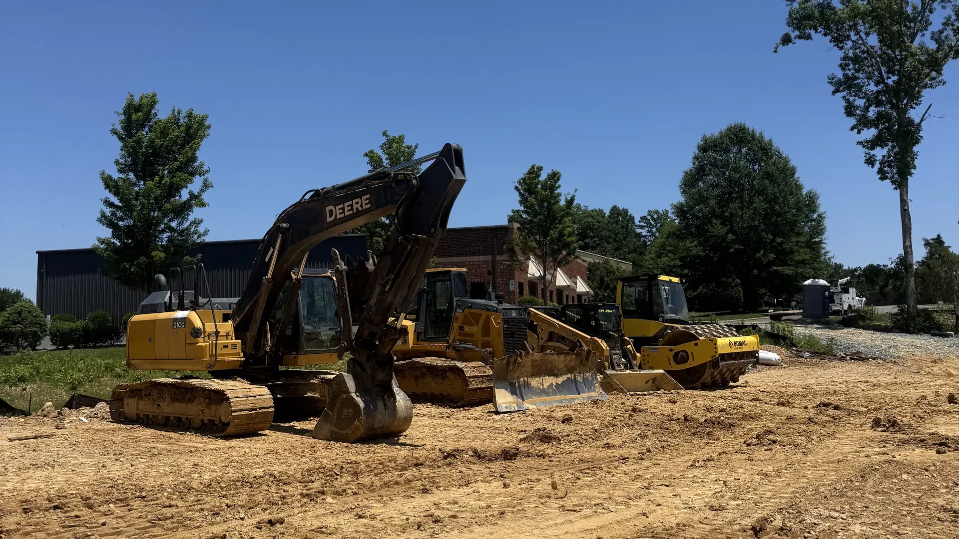 Excavator, dozer, roller and track loader parked on a graded red clay pad under a clear blue sky at a commercial site east of Charlotte