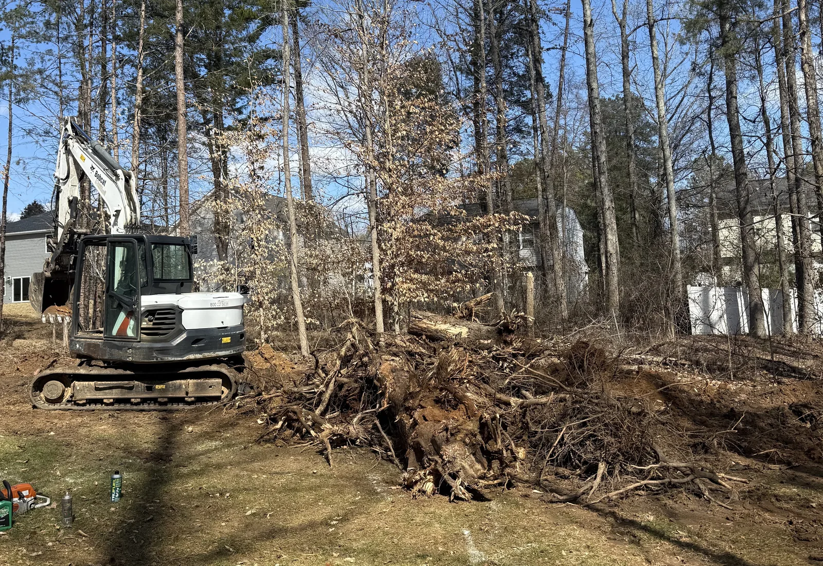 Excavator beside a pile of pulled stumps and brush on a cleared residential lot