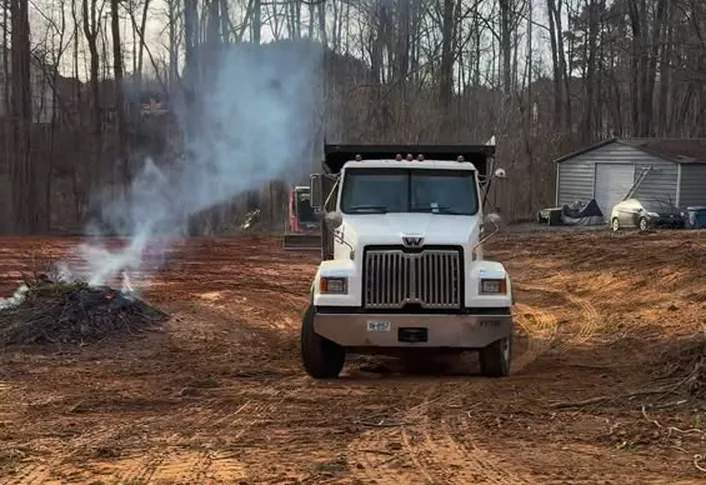 Dump truck parked on a cleared red clay lot beside a smoking brush pile