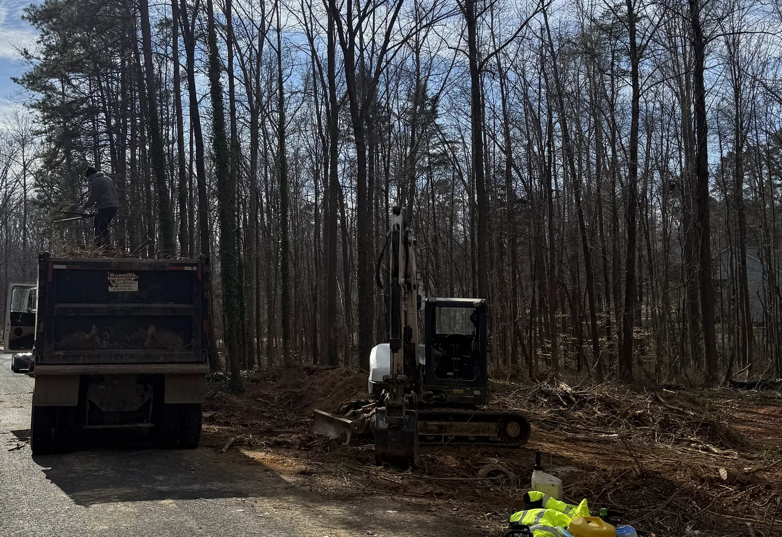 Brush being loaded into a dump truck beside an excavator at a wooded lot clearing