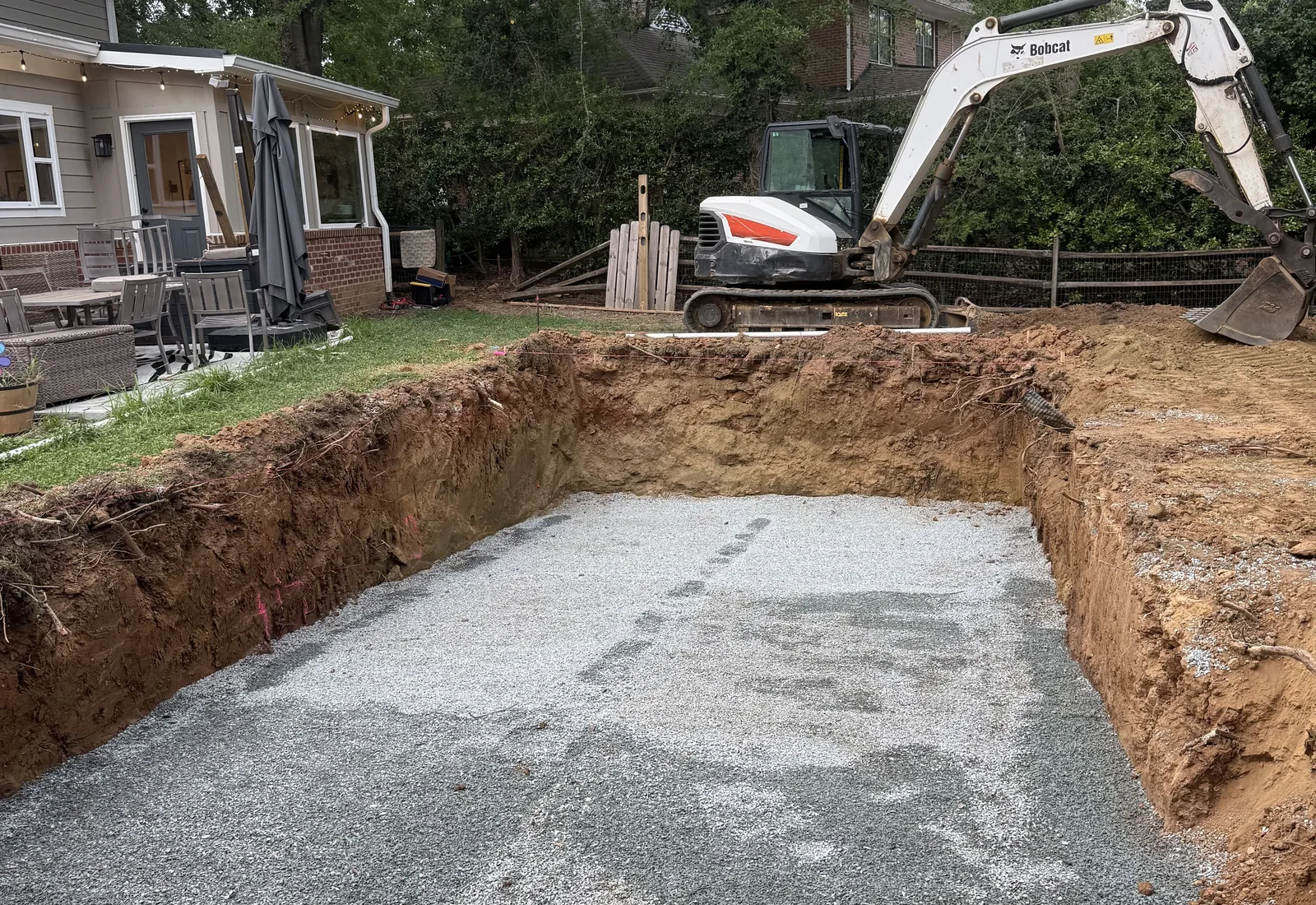 Pool excavation dug to a rectangle in a backyard and based with gravel, a mini excavator parked at the far edge