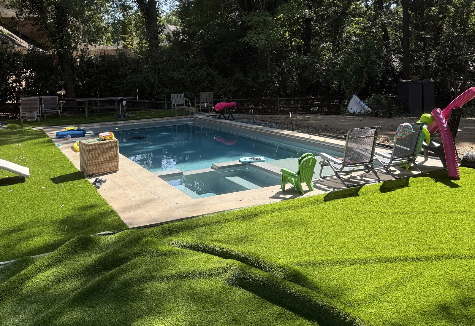 Finished inground pool with turf, patio chairs and a slide in a backyard shaded by tall trees