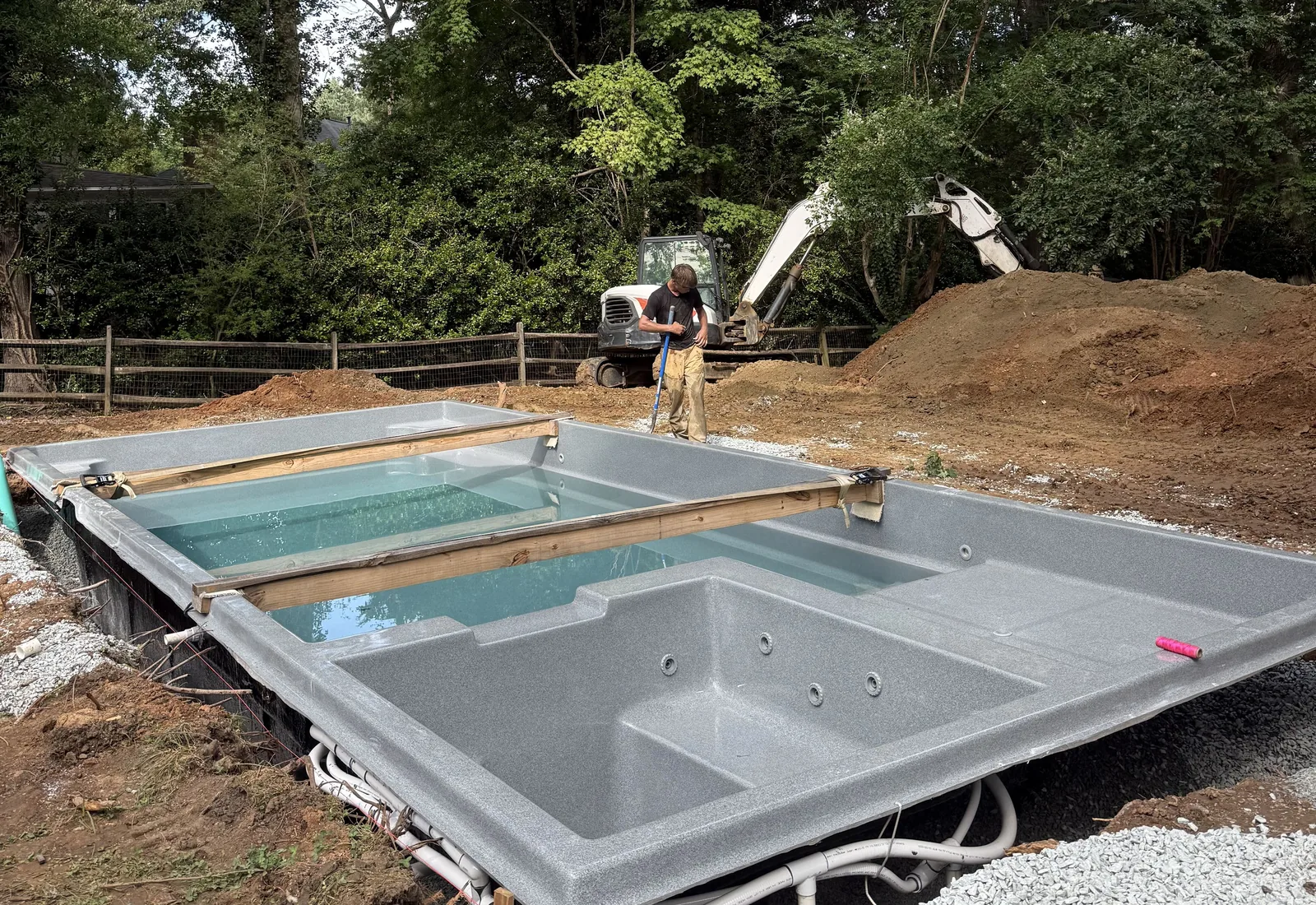 Fiberglass pool shell set on its gravel base in a backyard dig, a crew member raking beside it and a mini excavator behind the spoil pile