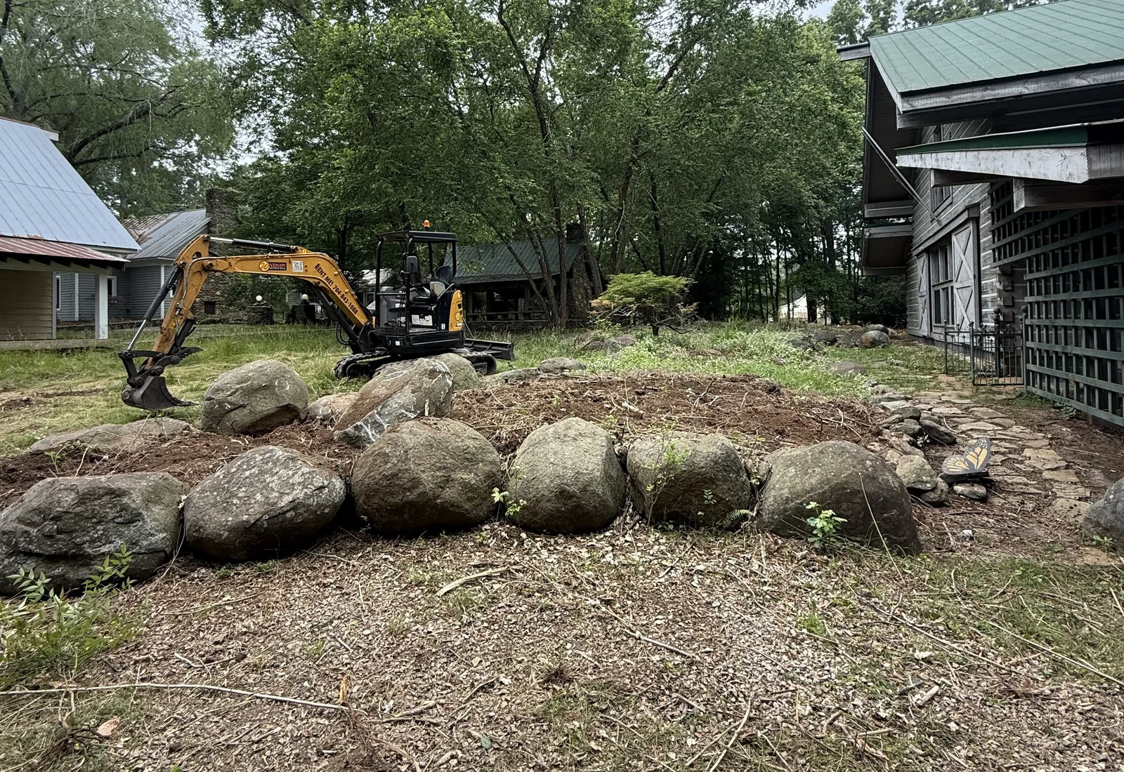 Mini excavator setting large boulders into a low rock wall along a slope beside a building