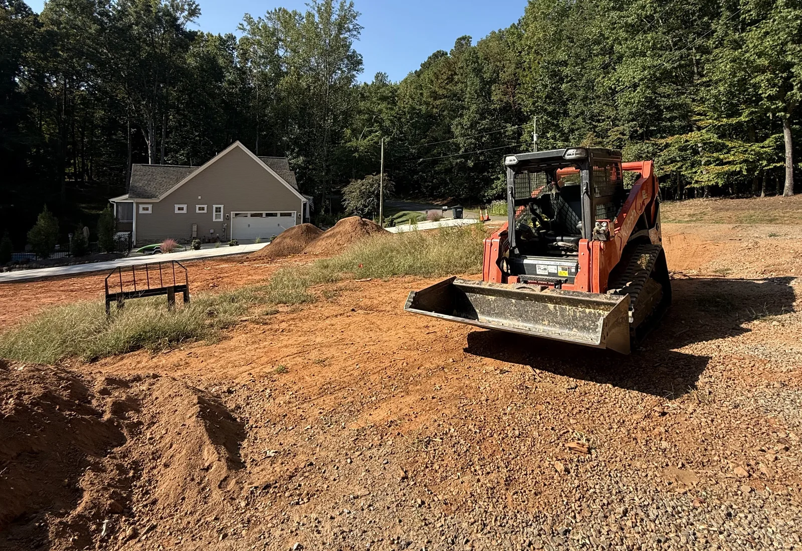 Track loader cutting into a red clay bank beside a house, the slope opened up and shaped