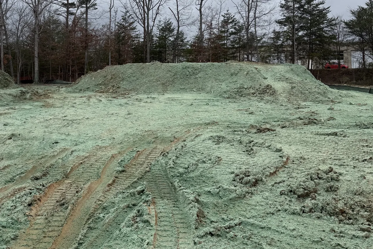 Dirt stockpile and graded ground covered in green seed and mulch on a construction site, tire tracks in the foreground