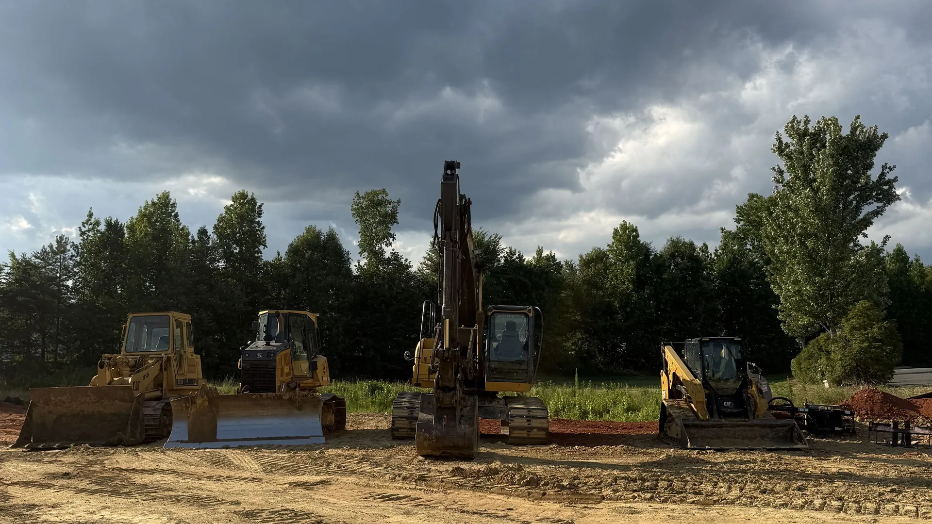 Our work: a track loader, a dozer, an excavator and a compact track loader lined up on a graded commercial pad under storm clouds