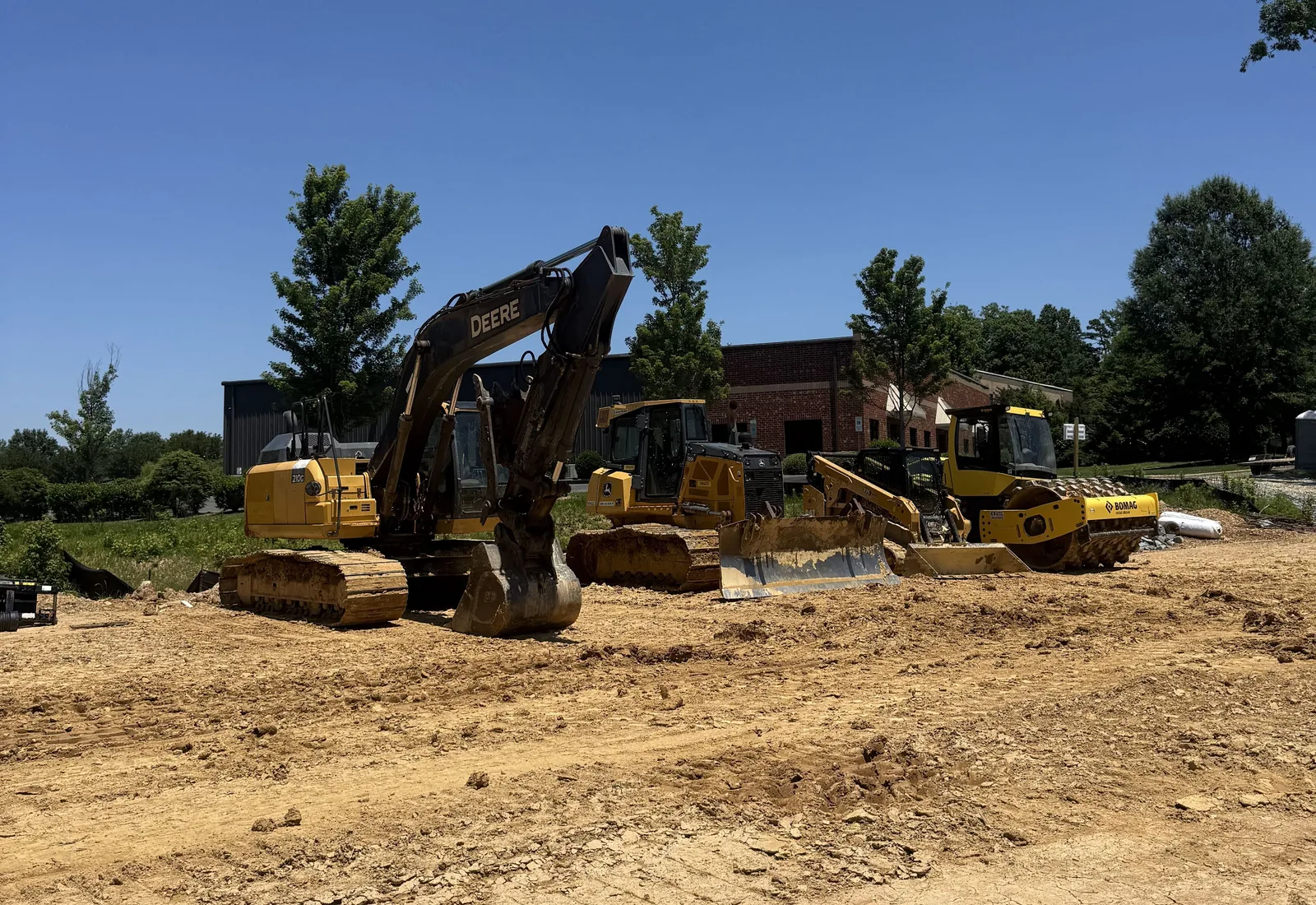 Excavator, dozer, compact track loader and a roller parked on a graded lot beside a brick commercial building