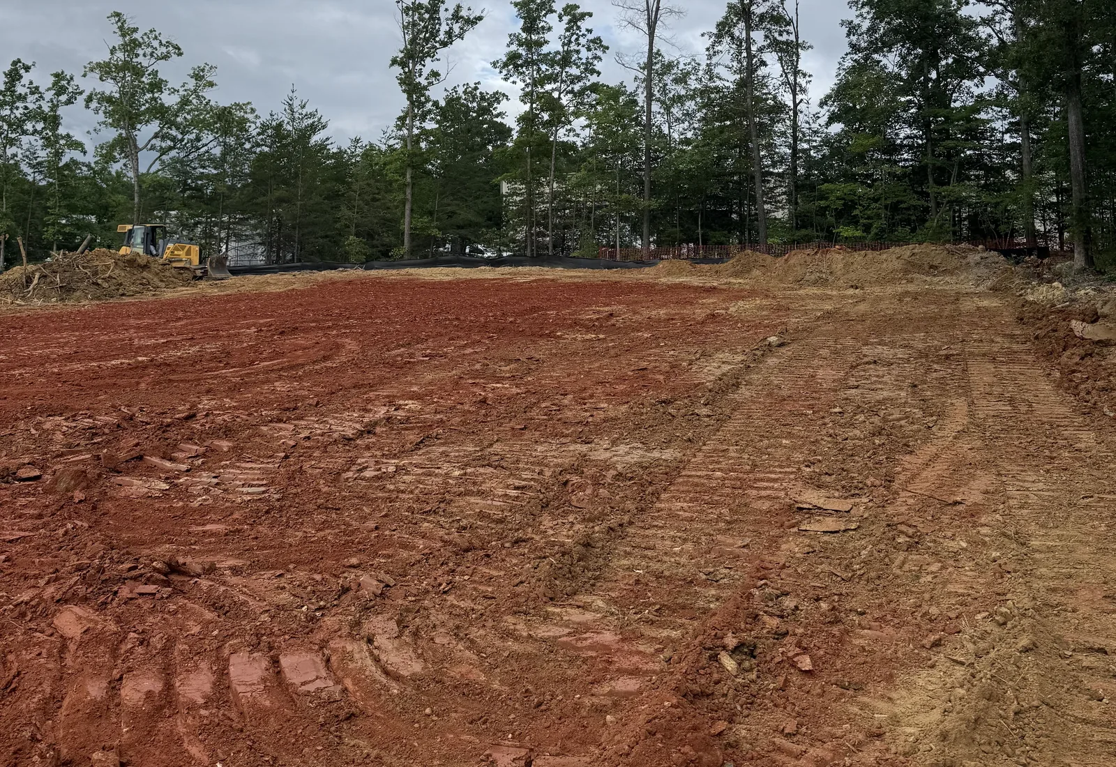 Graded red clay pad with silt fence and orange safety fence along the tree line and a dozer at the far edge
