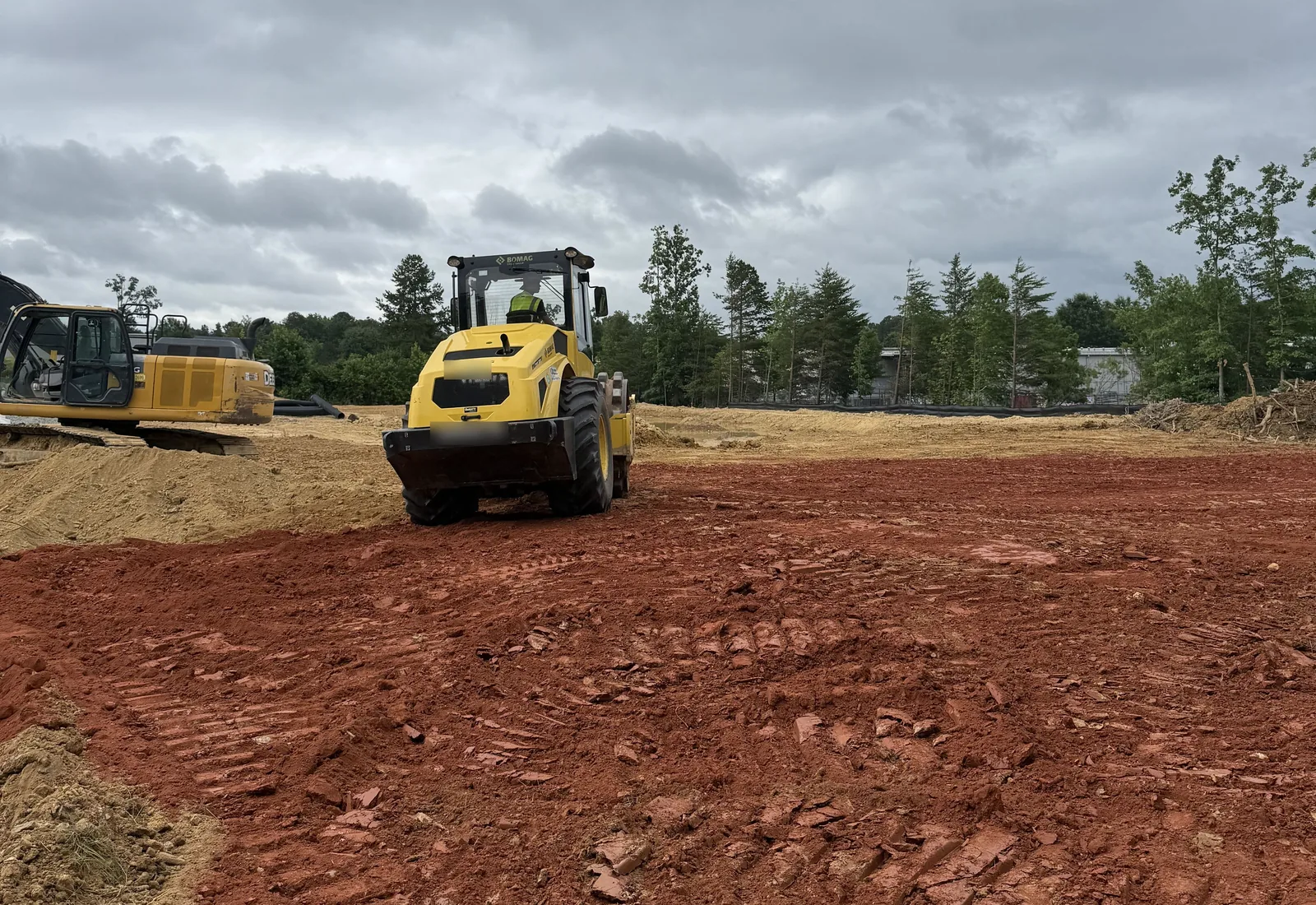 Vibratory roller compacting a red clay building pad with an excavator parked at the edge