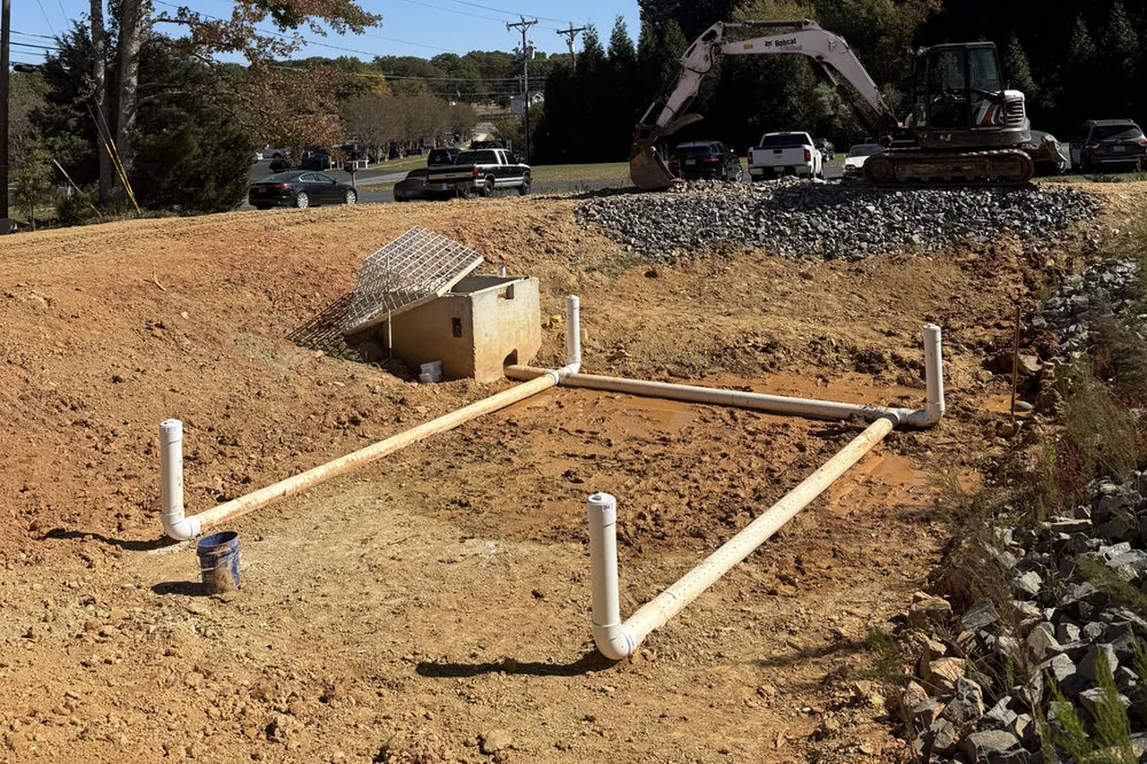 White PVC pipe runs laid out on a graded pad beside a small concrete box, with excavators parked behind