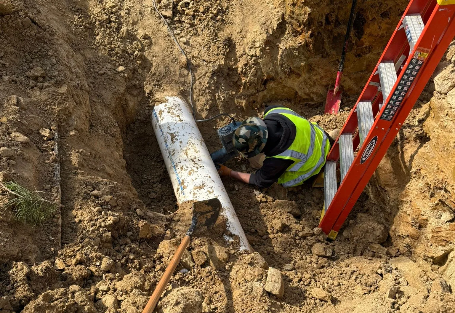 Open trench cut in clay with a large white pipe at the bottom, a crew member at work and a ladder set against the trench wall
