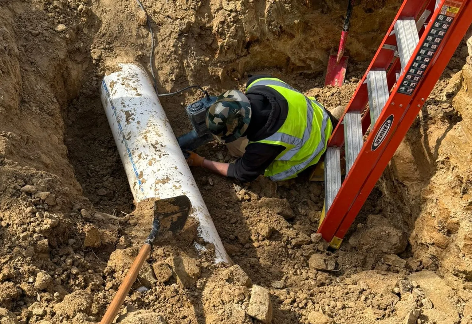 Crew member in a hi-vis vest working beside a large white pipe at the bottom of an open trench in clay