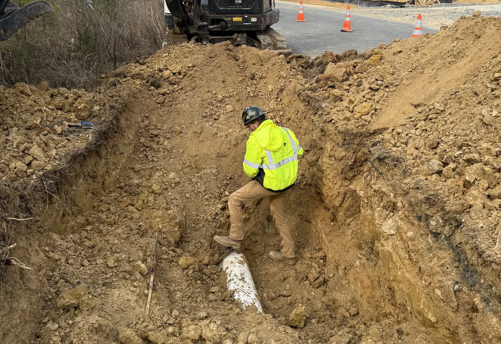 Deep trench cut in red clay at a commercial site with an excavator working above it