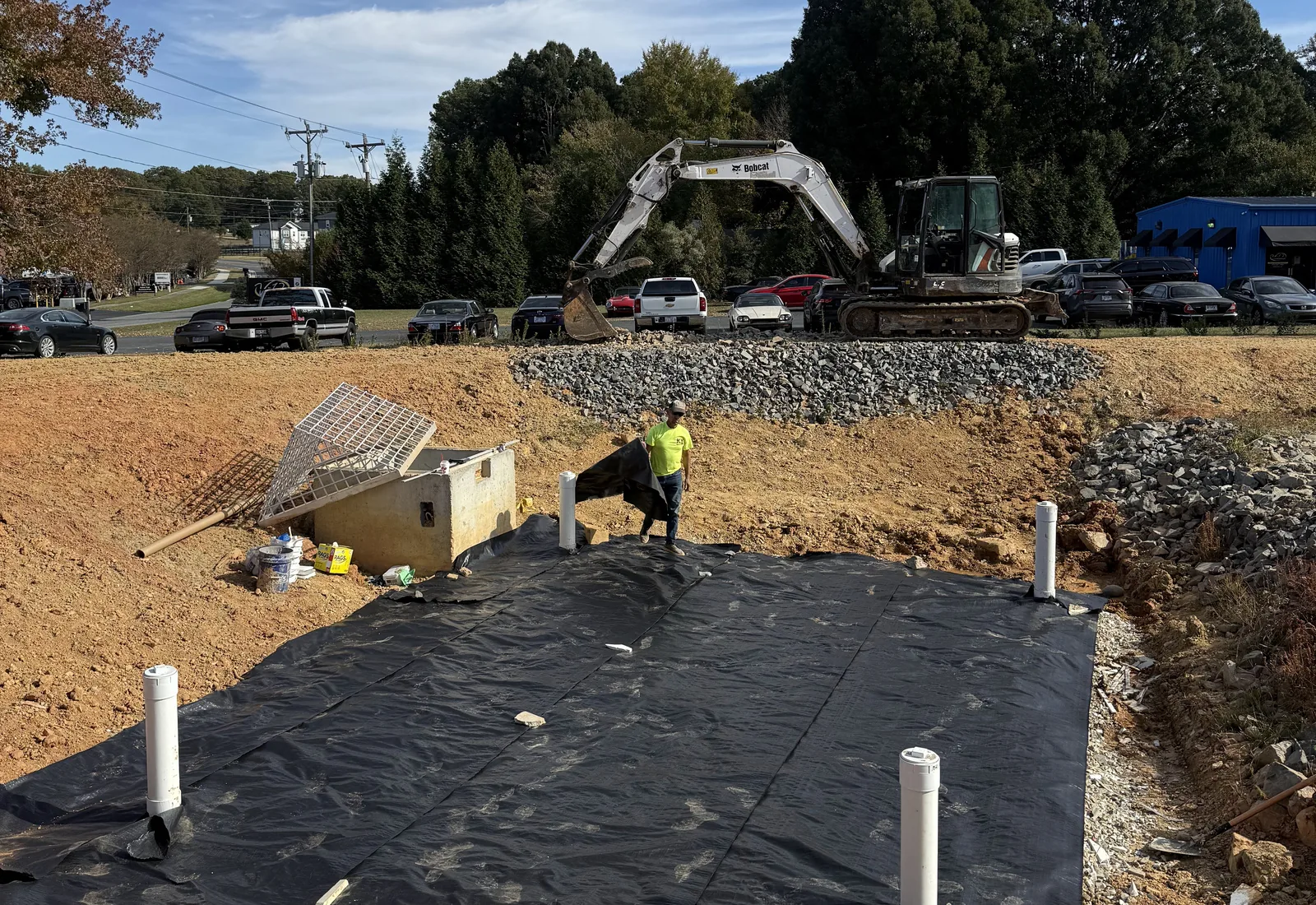 Gravel bed with pipe risers laid over liner in an excavation, excavator and stone stockpile behind