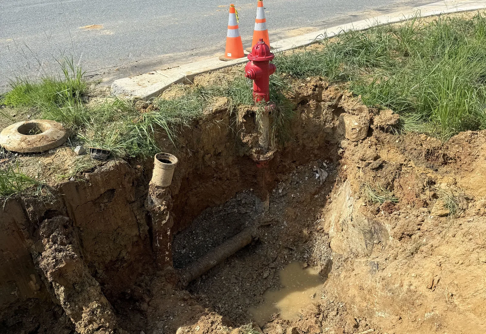 Excavation around a water main and valve at a new fire hydrant beside a road, cones set on the pavement