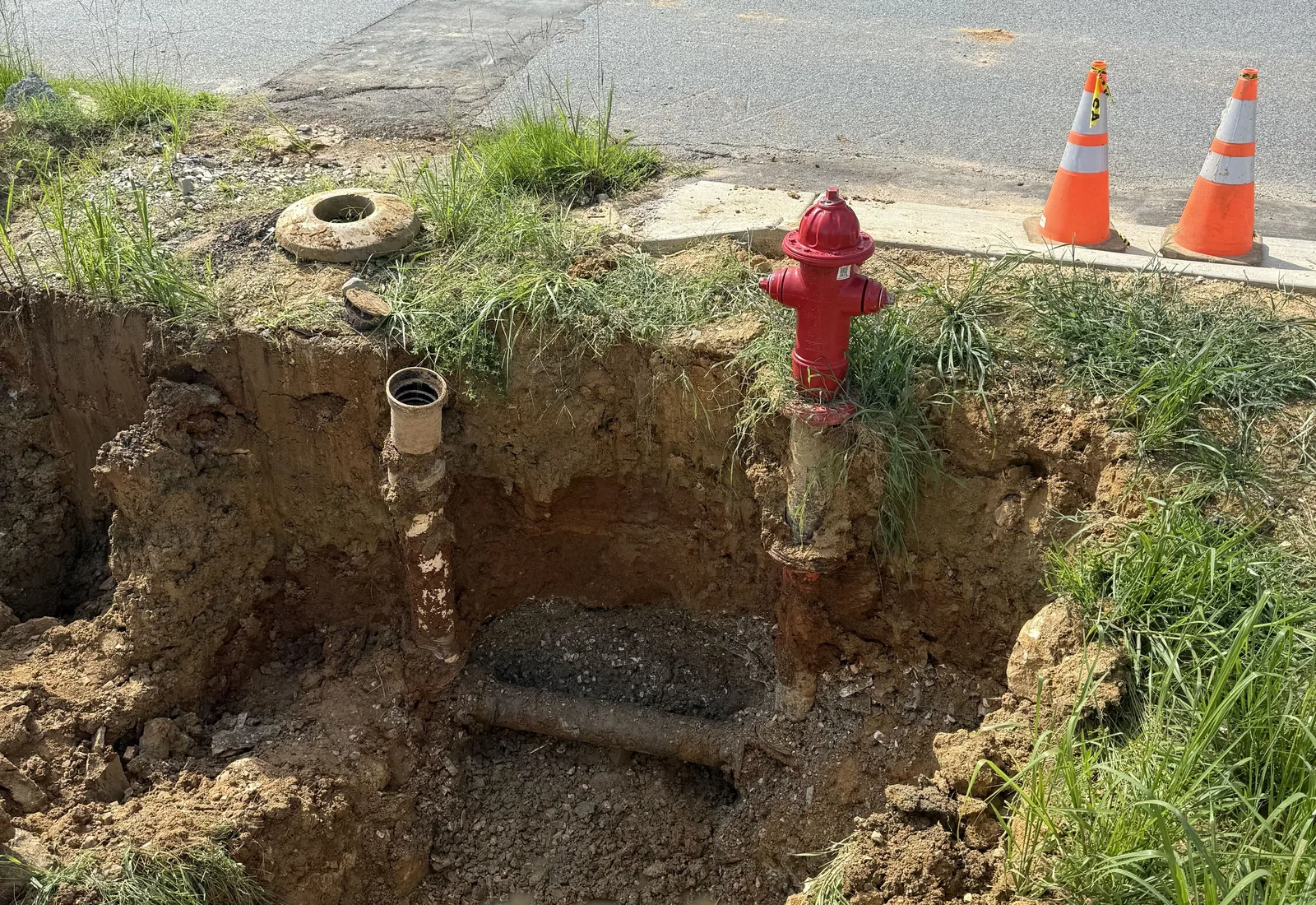 Open excavation beside a road exposing a water main and valve box next to a new fire hydrant, cones on the pavement