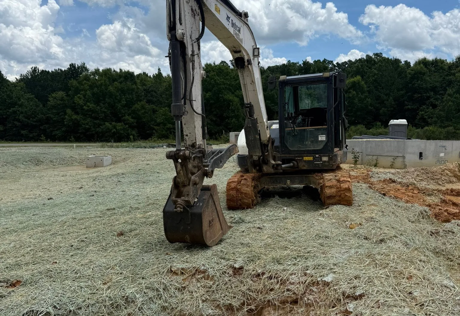 Excavator beside a concrete storm structure set in an open excavation on a straw covered site